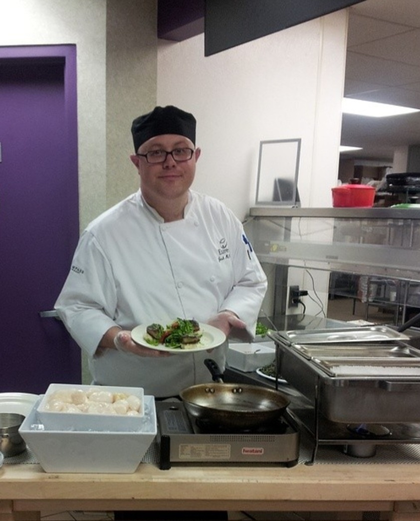 Joshua McCoury working in a kitchen wearing a white chef coat and smiling for the photo while holding a plate of food